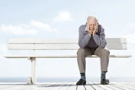 An elderly man sitting alone on a bench, resting his head on his hands with a thoughtful expression, against a bright sky and ocean background.